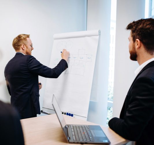 Exit Strategie Zwei Männer in Anzügen befinden sich in einem modernen Büro. Ein Mann steht und schreibt auf einem großen Flipchart, während der andere an einem Tisch mit offenem Laptop sitzt und aufmerksam zuschaut.